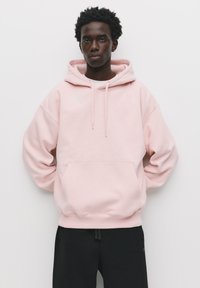 Helles rosa Kapuzensweatshirt mit einer Vordertasche, weichem Material und Kordelzügen. Darunter werden schwarze Hosen getragen.