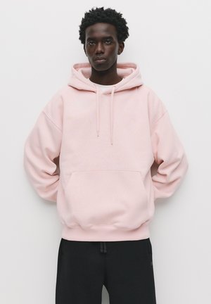 Sweat-shirt à capuche rose clair avec une poche kangourou à l'avant, texture douce et cordons de serrage. Un pantalon noir est porté en dessous.