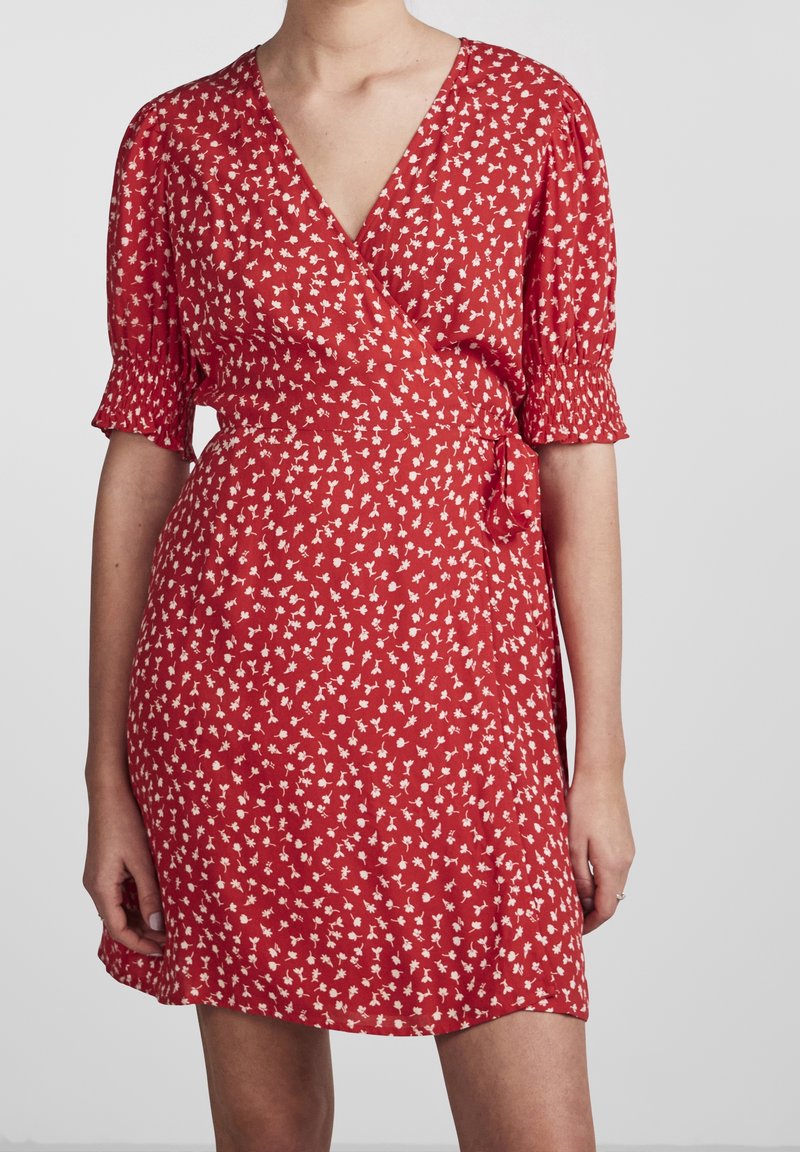 Robe portefeuille rouge avec petit motif floral blanc, manches courtes avec poignets froncés, portée par une personne debout sur fond uni.