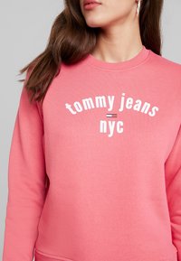 Rosa sweatshirt med texten "tommy jeans = nyc" i vitt. Rund hals, långa ärmar, mjukt material. Minimalistisk design med logotypdetalj.