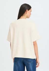 T-shirt beige oversize à manches courtes, en tissu doux. Il présente un col rond et une texture subtile, associé à un jean en denim bleu foncé.