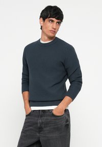 Dark blue ribbed sweater with a round neckline, long sleeves, and a fitted hem, paired with gray denim jeans. Textured fabric detail visible.