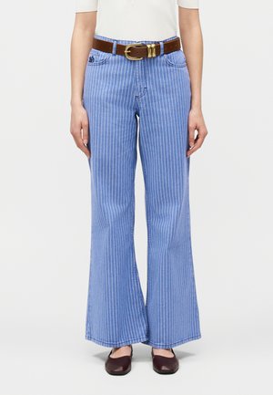 Osoba ubrana w niebieskie, szerokie spodnie z wysokim stanem w cienkie paski, z brązowym skórzanym paskiem i ciemnobrązowymi balerinami, stojąca na białym tle.