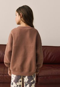 Hnedá oversized mikina s padnutými ramenami, rebrovaným golierom a textúrovanou látkou; kombinovaná s bielymi nohavicami s čiernym vzorom kravských škvŕn.