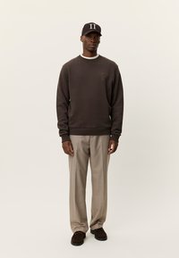 Καφέ φούτερ με υφασμάτινη λεπτομέρεια στην τσέπη, συνδυασμένο με μπεζ καρό παντελόνι. Το μοντέλο φορά σκούρο καπέλο με λευκές γράμματα και καφέ παπούτσια.