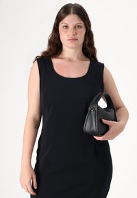Vestito nero con scollo a barchetta, texture liscia e design aderente. Tiene in mano una piccola borsa di pelle nera dalla forma arrotondata e con manico corto.