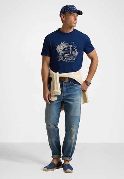 Marinblå bomullströja med grafik av en segelbåt, ljusbeige stickad tröja knuten i midjan, slitna blå jeans och blå espadrillos.