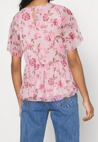Růžový květinový top s peplum střihem vyrobený z lehkého, průsvitného materiálu s krátkými rukávy. Obsahuje klíčovou díru na zádech a květinový vzor v odstínech růžové a zelené.