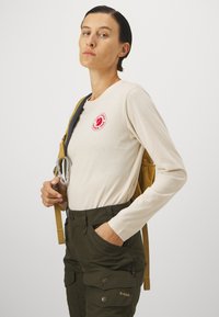 Pika diseiniga beež pikkade varrukatega särk punase ümmarguse logoga, millele on paariks roheline kamuflaazipüksid taskutega ja kollane seljakott rihmadega.