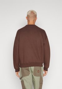 Brązowy, oversize sweter z okrągłym dekoltem i prążkowanymi mankietami, zestawiony z zielonymi spodniami cargo z patchworkowym wzorem i logo na tylnej kieszeni.
