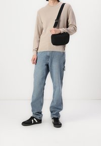 Persoon met een beige trui, lichtblauwe jeans, zwarte sneakers met witte strepen, en een zwarte schoudertas voor een witte achtergrond.