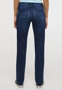 Osoba oblečená v tmavě modrých rovně střižených džínách a modrých teniskách se textem "MUSTANG", stojící na jednolitém světlém pozadí.