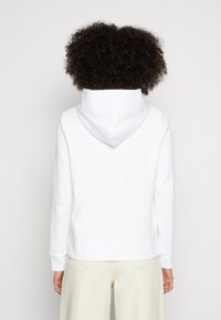 Sudadera blanca con capucha, puños y dobladillo acanalados. Textura suave, diseño simple. Combinada con pantalones de color claro.