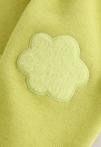Tissu jaune clair avec un patch de fleur en peluche verte cousu avec des bords festonnés, sur un fond de texture similaire.