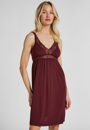 Blonde vrouw die een mouwloze bordeauxrode nachtkleding draagt met kanten rand langs de halslijn en onder de borst, staand tegen een effen achtergrond.