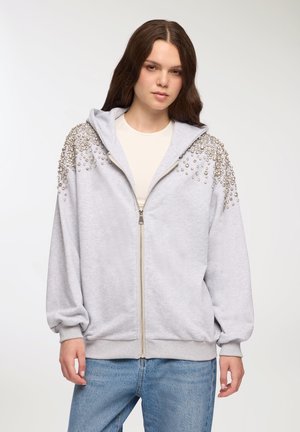 Felpa grigia con zip e cappuccio, decorata con perline argentate sulle spalle, vestibilità ampia e polsini e orlo a coste. Indossata sopra una maglietta chiara.