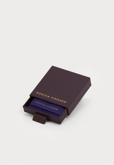 Scatola per gioielli rettangolare viola con il logo dorato "MONICA VINADER", parzialmente aperta per rivelare una scheda blu all'interno con testo e cuciture abbinate.