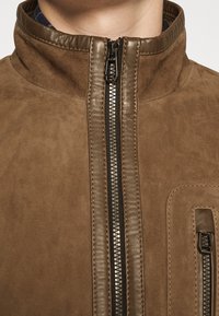 Brun jacka med hög krage, kontrasterande läderdetaljer och dragkedjestängning framtill med sidofickor.