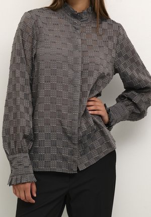 Kobieta ubrana w szarą teksturowaną bluzkę z długim rękawem i wysokim kołnierzem oraz czarne spodnie, pozująca z jedną ręką na biodrze.