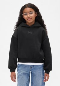 Melnas kapuces džemperis ar GAP logotipu priekšpusē. Izceļas ar platām piedurknēm un īsu rāvējdatoru dizainu, kopā ar gaiši zilām džinsa biksēm. Gluda auduma tekstūra.