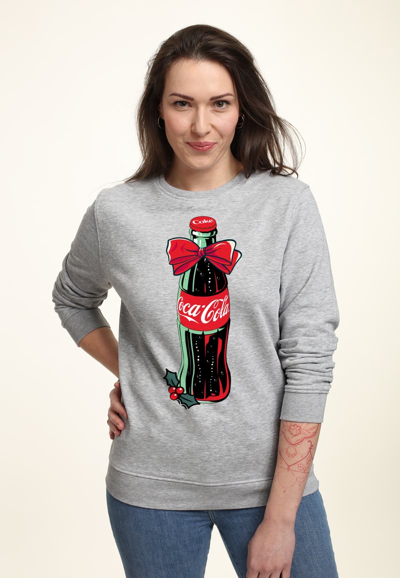 Felpa grigia con una grafica di una bottiglia di Coca-Cola decorata con un fiocco rosso e accenti di agrifoglio, su uno sfondo chiaro.