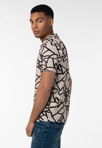 Beige Poloshirt mit kurzen Ärmeln und einem schwarzen abstrakten Muster. Das Material wirkt weich, hat eine lässige Passform und ein klassisches Kragen Design.