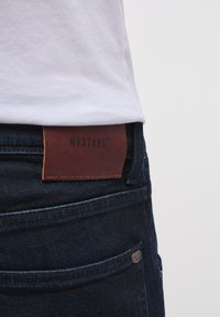 Jeans in denim blu scuro con un'etichetta in pelle marrone sulla vita etichettata "MUSTANG". Presentano tasche standard e dettagli in metallo.