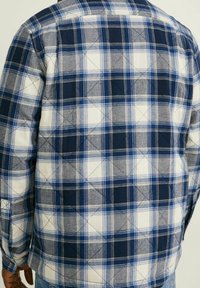 Rutigt skjorta med blå, marinblå och vita färger. Har en vadderad textur och knäppta ärmslut. Tvåfärgat rutmönster över ryggen.