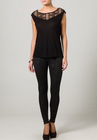 Musta paita, jossa on pitsinen kaula-aukko ja lyhyet hihat; yhdistetty korkeavyötäröisiin mustiin legginsseihin, joissa on monimutkainen teksturoitu kuvio.