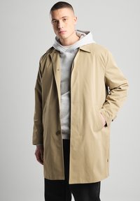 Beige trench coat with a relaxed fit, button closure, side pockets, and a smooth texture, layered over a grey hooded sweatshirt.