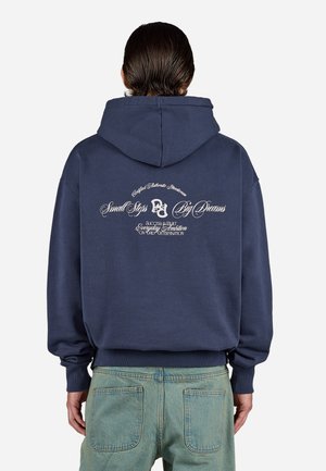 Person with short, slicked-back hair wearing navy hoodie with white text reading "Small Steps Big Dreams" and faded blue jeans, seen from behind.