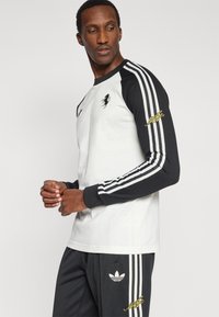 Αθλητικό τοπ σε ασπρόμαυρο με μακριά μανίκια, τρεις ρίγες στα μανίκια και λογότυπο στο στήθος· συνοδεύεται από μαύρο παντελόνι με λογότυπο.