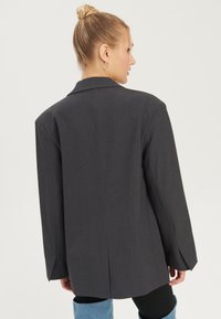 Frau mit blondem Haar im Dutt trägt einen dunkelgrauen übergroßen Blazer, schwarze Leggings und hellblaue kniehohe Stiefel, seitlich gewandt.