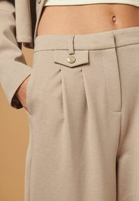 Beige tailleurhousut, joissa on etupleikkeitä, koristeellinen läppä ja nappikiinnitys sekä sivutaskut pehmeästä ja sileästä kankaasta.