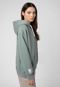 Lichtgroene hoodie van zacht materiaal, met een ritssluiting aan de voorkant, verstelbare trekkoordjes, zijzakken en een logo-opdruk op de mouw.