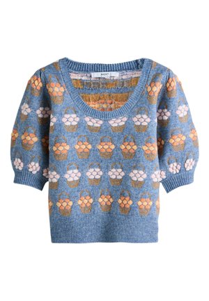 Femme portant un pull bleu à manches bouffantes à fleurs, un pantalon large gris clair, et tenant un petit sac à main mauve.