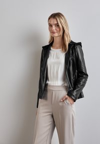 Giacca in pelle nera con cappuccio, chiusura con zip e tasche laterali, indossata sopra una camicetta bianca e pantaloni beige chiaro. Texture morbida.