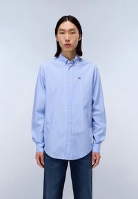 Light blue button-up shirt with vertical white stripes, featuring a pointed collar and small logo detail on the chest. Worn with dark jeans.