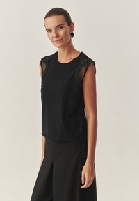 Fekete, ujjatlan felső, hálós panel díszítéssel a vállakon, klasszikus kerek nyakkivágással és sima, texturált felülettel.