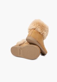 Tanfarvede ankelstøvler i ruskind med pelskant og strukturerede gummisåler. Har en forhøjet hæl for støtte og syningsdetaljer på siderne.