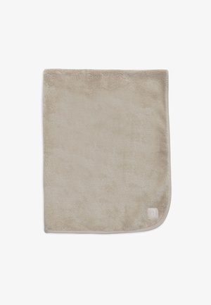 Zachte, beige pluche deken met een gladde textuur en ronde hoeken. Bevat een subtiele logo in de onderste hoek.