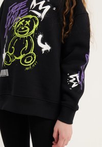 Sweatshirt preto com gráficos em estilo spray de tinta verde e branca, um urso cartoon e texto; punhos e barra canelados; corte descontraído.