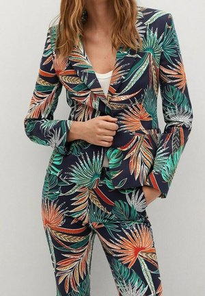 Femme portant un blazer cintré et un pantalon avec un motif coloré de feuilles tropicales en vert, orange et blanc sur un fond sombre.