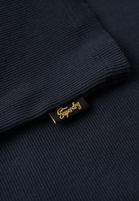 Tissu côtelé bleu marine avec une texture lisse ; présente une étiquette noire avec le texte doré "Superdry" sur le bord.
