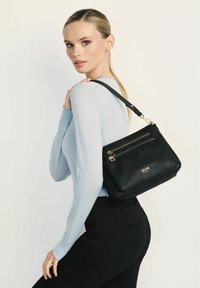 Czarna skórzana torebka z dwoma przednimi suwakami, teksturowana powierzchnia i akcenty w kolorze złotym. Nosi się ją na ramieniu na jasnym tle.