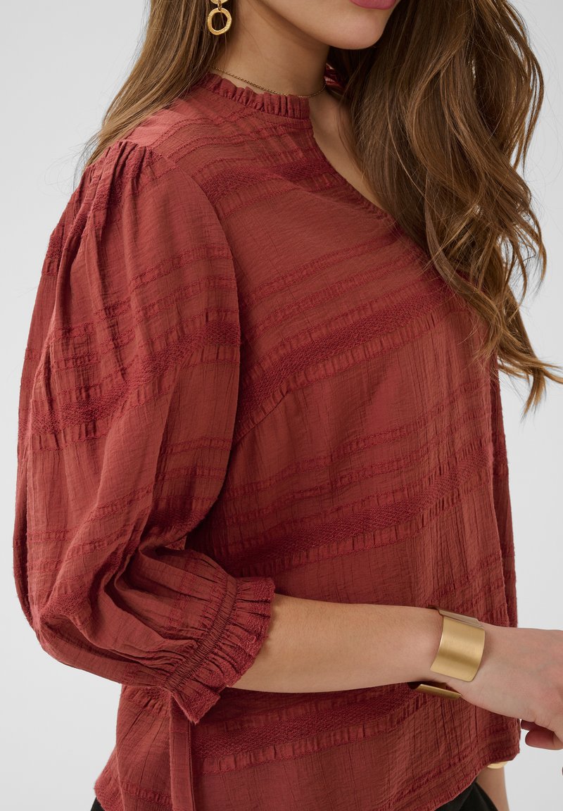 Femme portant un chemisier texturé rouge rouille à manches froncées, un bracelet manchette en or et des boucles d'oreilles géométriques en or, les cheveux lâchés sur l'épaule.
