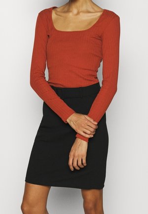 Persoon draagt een aansluitende baksteenrode lange mouwen top met een vierkante halslijn, gecombineerd met een hoge taille zwarte rok, armen losjes gekruist.