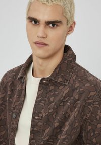 Veste en denim marron à motifs avec un col, dotée d'une texture douce et de boutons-pression. Portée sur une chemise de couleur claire.