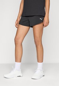 Person som bär svarta Puma träningstights, vita strumpor, och vita sneakers, står mot en ljus, enkel bakgrund.