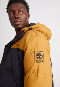 Pufferjakke i sort og gylden gul med hætte, lynlås og et Timberland-logo-patch på venstre ærme. Glat tekstur.